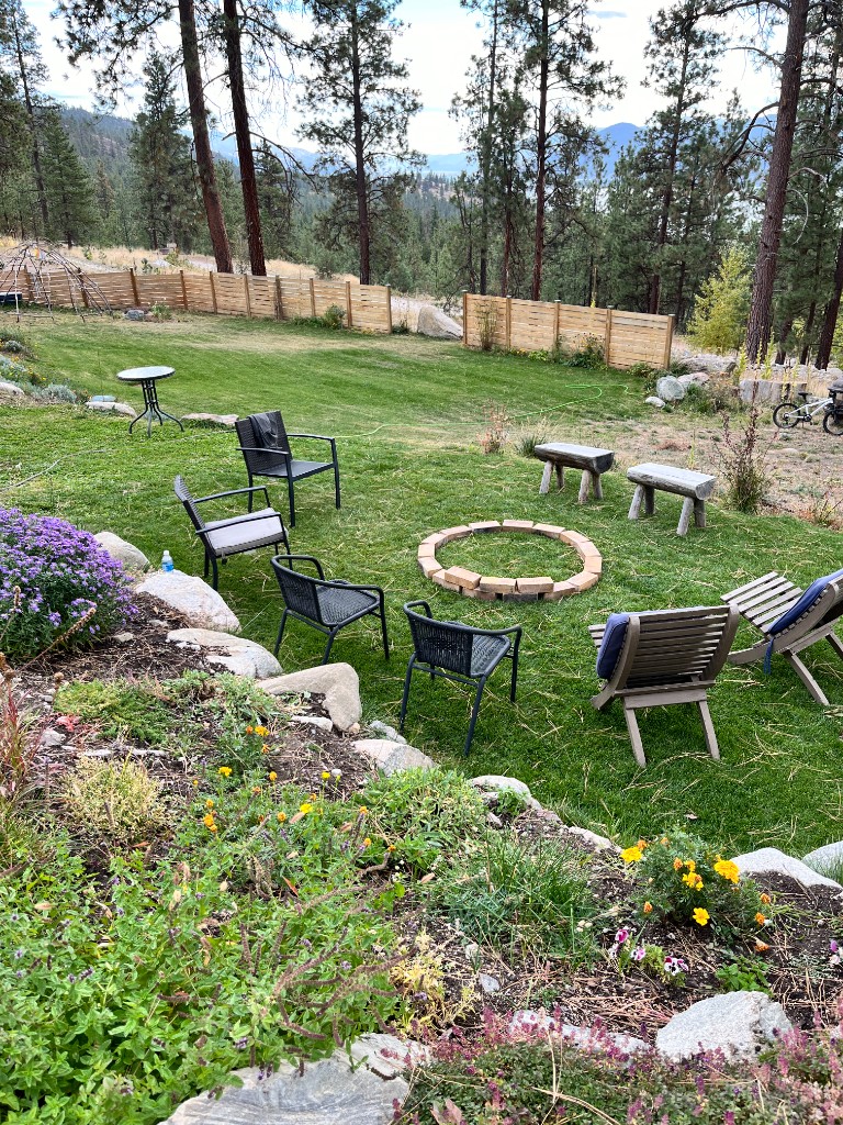 Brick fire pit with chairs, garden beds, and a lake glimpse through the trees